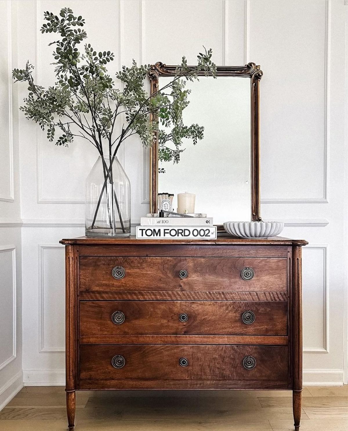 Cómoda vintage decorado con un espejo y unas ramas con hojas verdes.