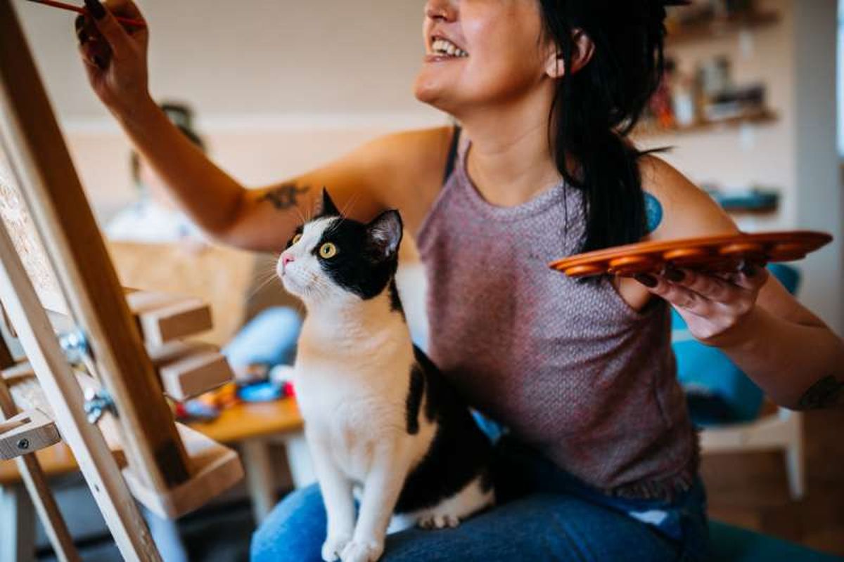 Mujer sonriente pintando un lienzo con un gato sobre las piernas.